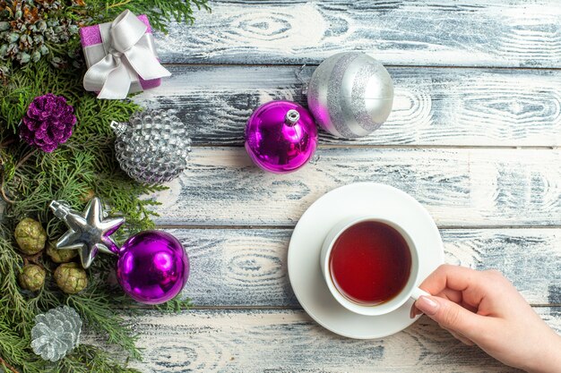 Vista superior de una taza de té en mano femenina regalo pequeño ramas de abeto juguetes de Navidad sobre fondo de madera