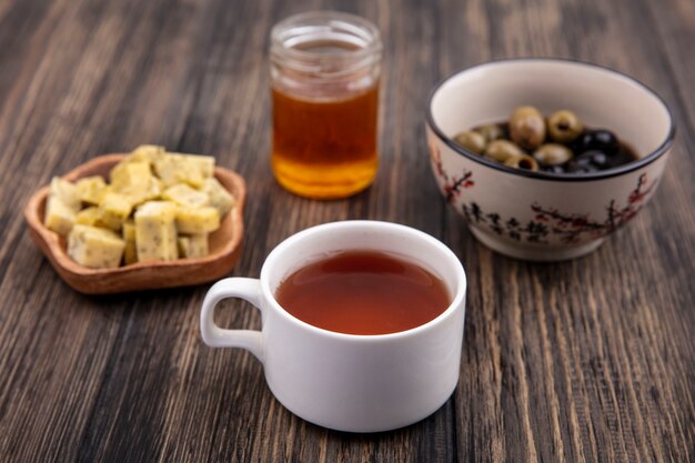 Vista superior de una taza de té con miel con aceitunas y rodajas de queso picado sobre un fondo de madera