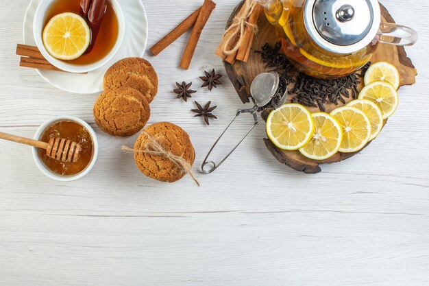 Vista superior de una taza de té negro galletas miel y té de hierbas en una olla de vidrio y limones de canela limón picados en una bandeja de madera sobre fondo blanco.