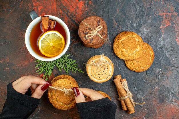 Vista superior de la taza de té palitos de canela diferentes galletas atadas con una cuerda en la mano femenina en la mesa de color rojo oscuro