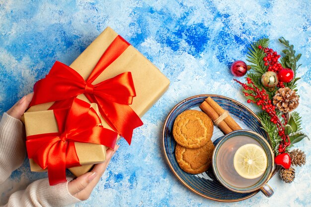 Vista superior de la taza de té, palitos de canela, galletas en platillo, regalos en mano femenina en la mesa azul