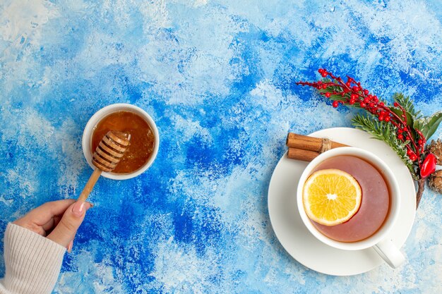 Vista superior taza de té palo de miel en mano femenina rama de árbol de Navidad en mesa azul con espacio de copia