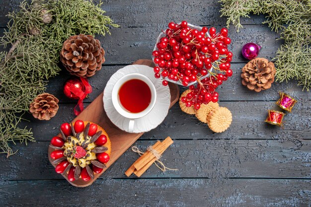 Vista superior de una taza de té y pastel de bayas en un plato de madera de grosella en un vidrio piñas juguetes de navidad hojas de abeto en la mesa de madera oscura