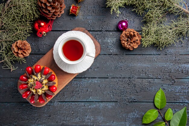 Vista superior de una taza de té y pastel de bayas en un plato de madera con piñas, juguetes navideños y hojas en la mesa de madera oscura.