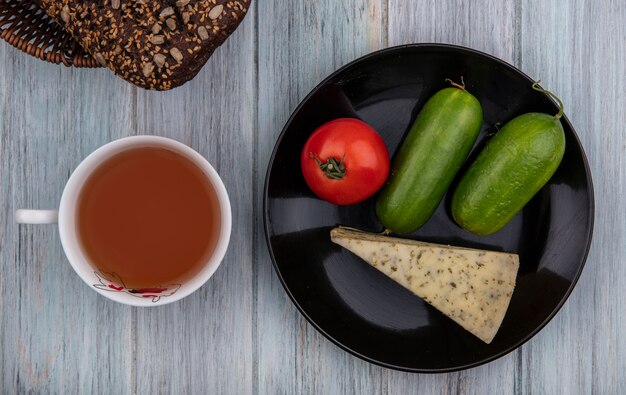 Vista superior de la taza de té con pepino de queso pan negro y tomate en un plato sobre fondo gris