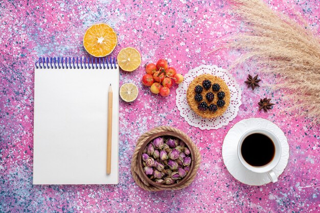 Vista superior de una taza de té con un pequeño pastel y un bloc de notas sobre el fondo rosa.