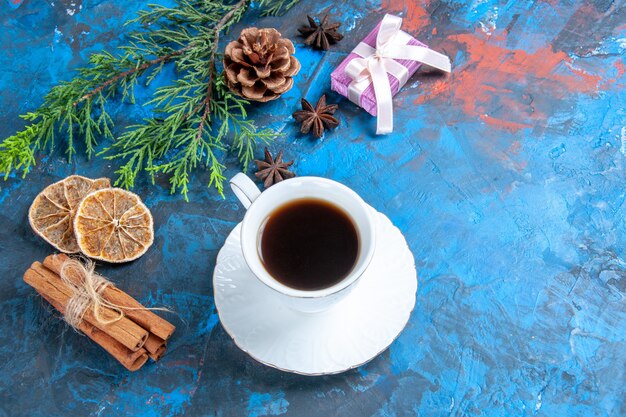 Vista superior de una taza de té, platillo blanco, ramas de pino, ramas de canela, anís, rodajas de limón secas sobre fondo azul.