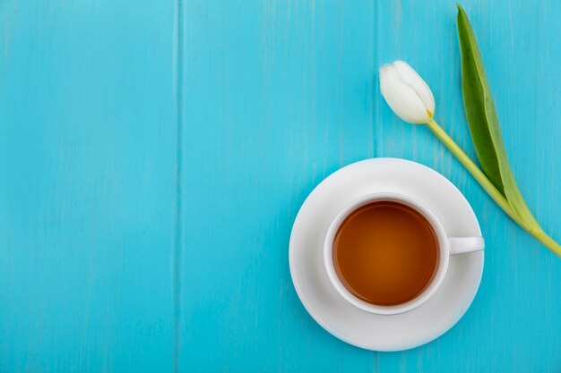 Vista superior de la taza de té en el platillo y flor sobre fondo azul con espacio de copia