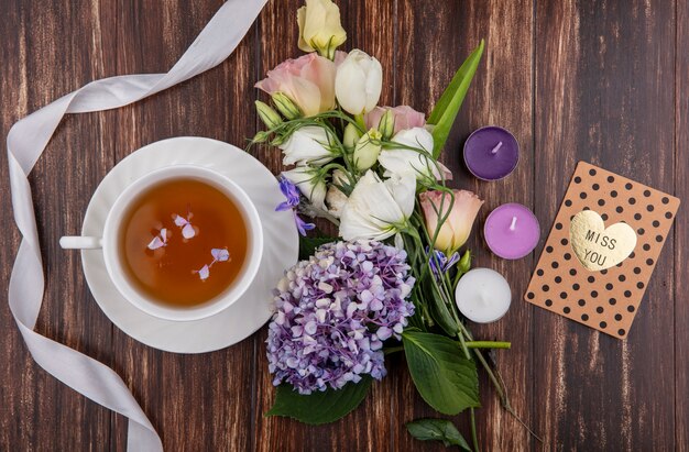 Vista superior de la taza de té en el platillo y flores con la cinta de la tarjeta miss you y velas sobre fondo de madera