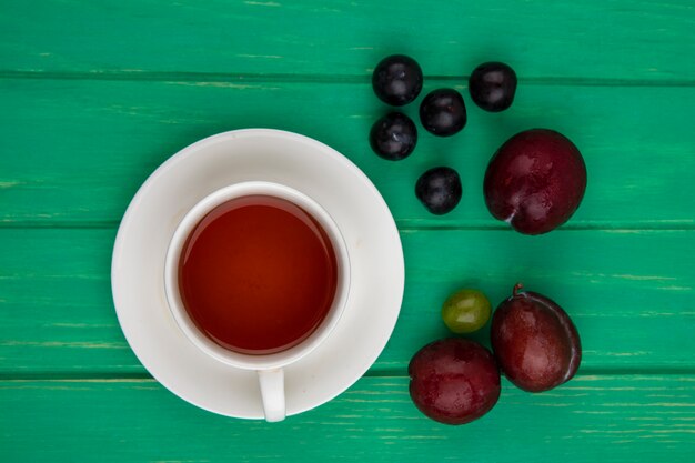 Vista superior de la taza de té en el platillo y el patrón de frutas como bayas de uva pluots y endrinas sobre fondo verde