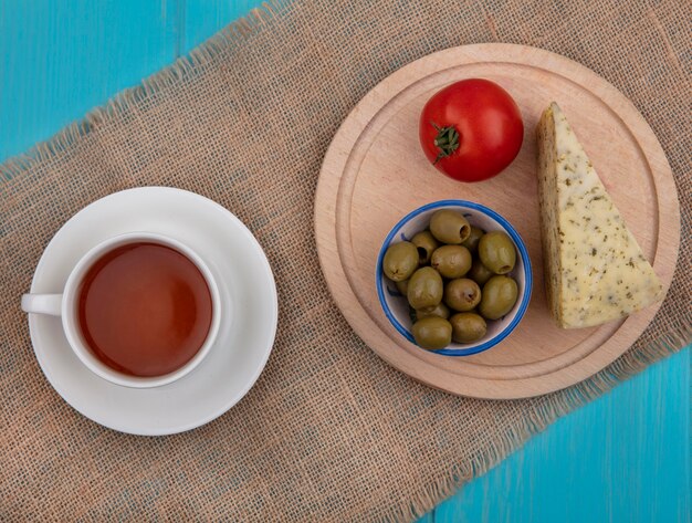 Vista superior de la taza de té con queso, aceitunas y tomate en un soporte sobre una servilleta beige