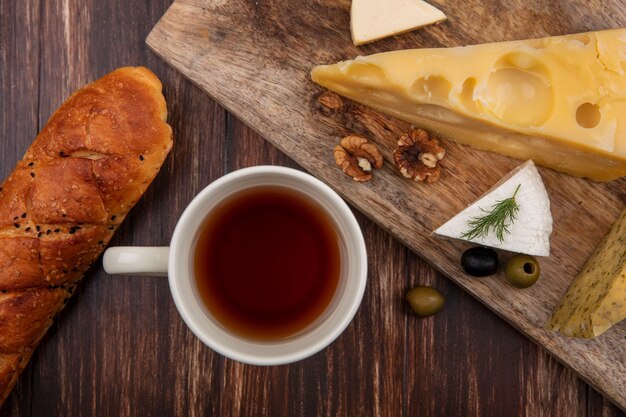 Vista superior de la taza de té con una rebanada de queso maasdam y queso feta con aceitunas en una tabla sobre un fondo de madera