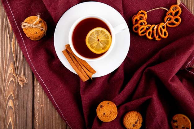 Vista superior taza de té con una rodaja de limón y canela con galletas sobre un mantel de color burdeos