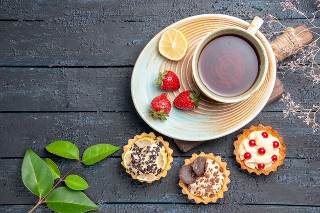 Vista superior de una taza de té, rodaja de limón y fresas en tartas de platillo deja en la mesa de madera oscura.