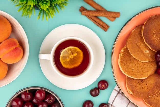 Vista superior de una taza de té con una rodaja de limón y un plato de panqueques con albaricoques y cerezas y canela sobre fondo azul.