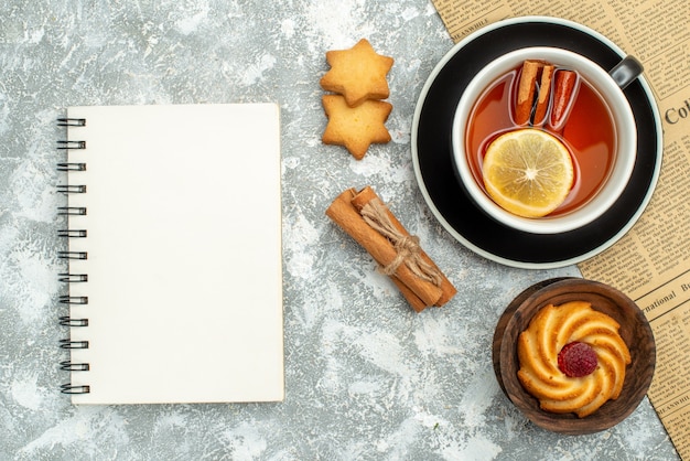 Vista superior de una taza de té con rodajas de limón y palitos de canela en galletas de cuaderno de periódico en superficie gris