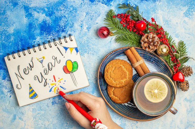 Vista superior de una taza de té, rodajas de limón, palitos de canela, galletas en el platillo, año nuevo escrito en el bloc de notas rojo pe en la mano de la mujer en el cuadro azul