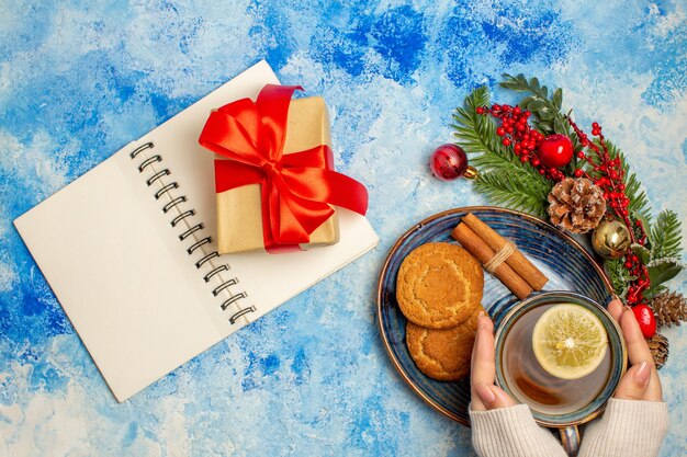 Vista superior de la taza de té, rodajas de limón, palitos de canela, galletas en el platillo, el bloc de notas, regalo de Navidad en la mesa azul