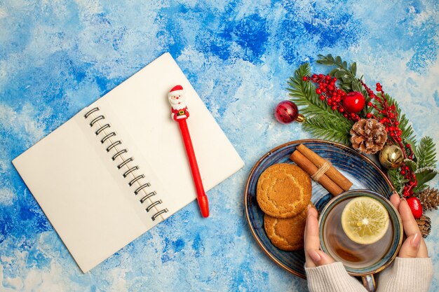 Vista superior de una taza de té, rodajas de limón, palitos de canela, galletas en un platillo, bolígrafo rojo en el bloc de notas en la mesa azul