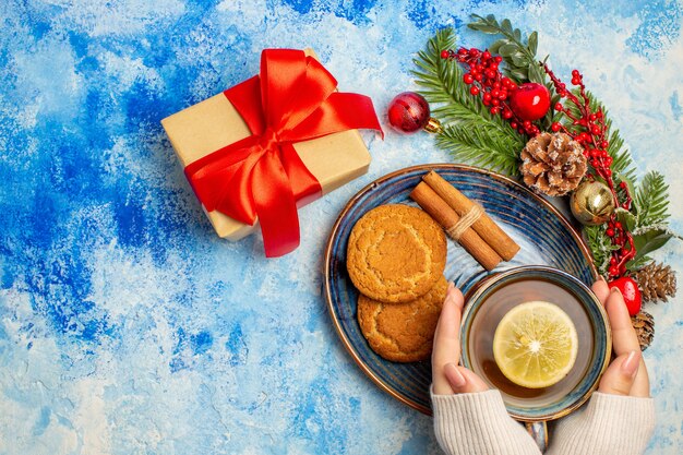 Vista superior de una taza de té, rodajas de limón, palitos de canela, galletas en platillo, regalo de Navidad en la mesa azul.