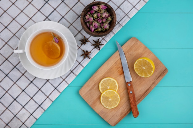 Vista superior de una taza de té con rodajas de limón en un tablero con un cuchillo con flores secas sobre un fondo azul claro