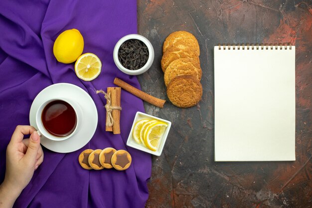 Vista superior de la taza de té, rodajas de limón en un tazón, canela en rama, té en un tazón en el bloc de notas de mantel morado en la mesa de color rojo oscuro