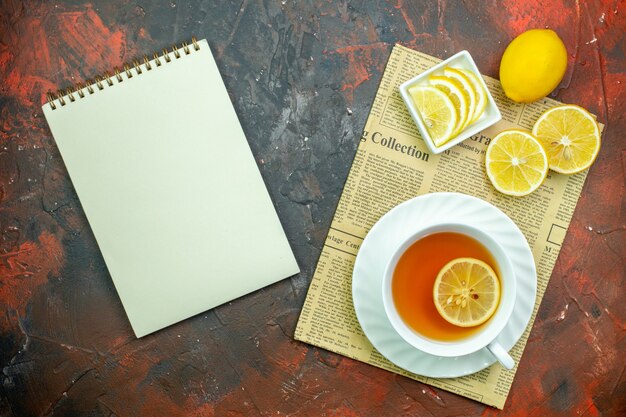 Vista superior de la taza de té de rodajas de limón en un tazón pequeño en un cuaderno de periódico sobre fondo de siena oscuro