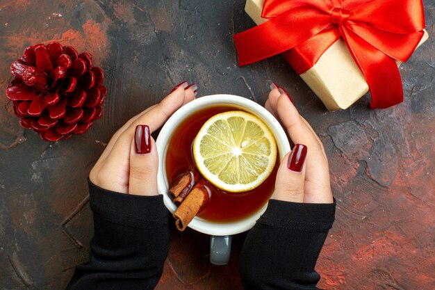 Vista superior de una taza de té con sabor a limón y canela en la mano femenina, regalo de Navidad, piña roja en la mesa de color rojo oscuro.