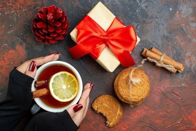 Vista superior de la taza de té con sabor a limón y canela en la mano femenina regalo de Navidad piña roja palitos de canela galletas en la mesa de color rojo oscuro