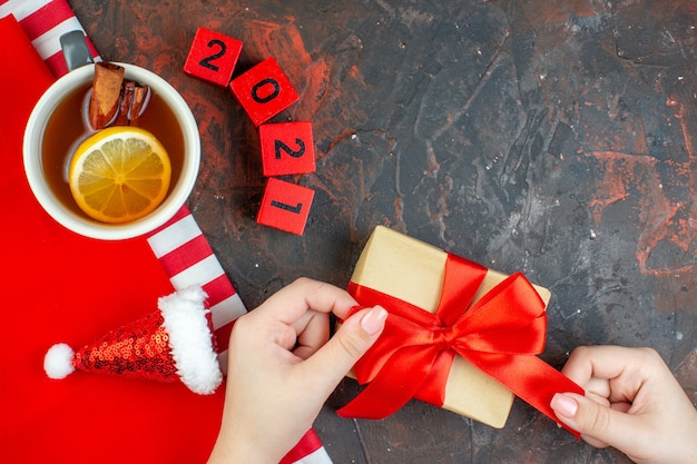 Vista superior taza de té con sabor a limón y canela mini gorro de santa sobre mantel rojo regalo de bloques de madera en mano femenina sobre mesa rojo oscuro