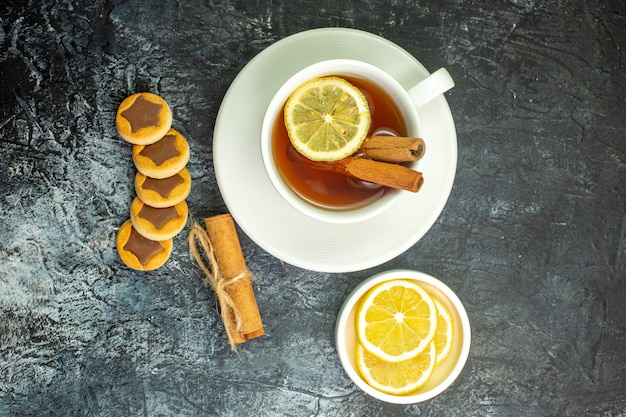 Vista superior de la taza de té con sabor a limón y canela rodajas de limón en pequeñas galletas de platillo palitos de canela en la mesa oscura