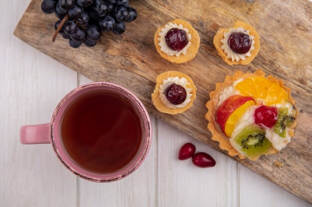 Vista superior de una taza de té con tartaletas y uvas negras sobre una tabla para cortar