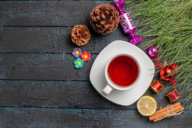 Vista superior de una taza de té una taza de té en el platillo blanco junto a las ramas de abeto limón canela con juguetes navideños y conos en el lado derecho de la mesa