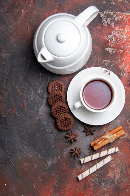 Vista superior de la taza de té con tetera y galletas en la mesa oscura