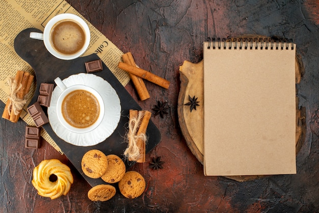 Vista superior de las tazas de café en la tabla de cortar de madera y un periódico viejo galletas canela limas barras de chocolate junto al cuaderno de espiral sobre fondo oscuro