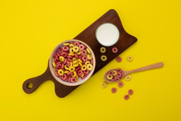 Vista superior del tazón de cereal y vaso de leche en la tabla de cortar con una cucharada de cereal sobre fondo amarillo