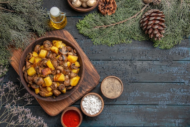 Vista superior desde un tazón de comida lejano de papas con champiñones en la tabla de cortar junto a las coloridas especias debajo del tazón de aceite de setas blancas y ramas de abeto