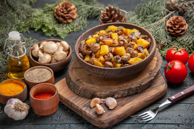 Vista superior del tazón de fuente de comida marrón tazón de papas con champiñones en la tabla de cortar junto al tenedor ajo especias coloridas aceite en botella y tazón de setas debajo de ramas con conos