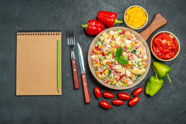 Vista superior del tazón de fuente de ensalada de verduras con cubiertos vegetales y bloc de notas en el lado de la mesa gris oscuro