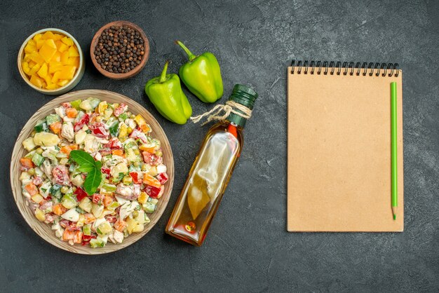 Vista superior del tazón de fuente de ensalada de verduras con tazones de fuente de pimiento y verduras botella de aceite pimientos y bloc de notas en el lado de la mesa oscura