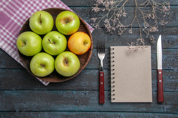 Foto gratuita vista superior del tazón de fuente de manzanas de siete manzanas de color amarillo verdoso sobre un mantel a cuadros junto a ramas, tenedor, cuchillo y cuaderno gris