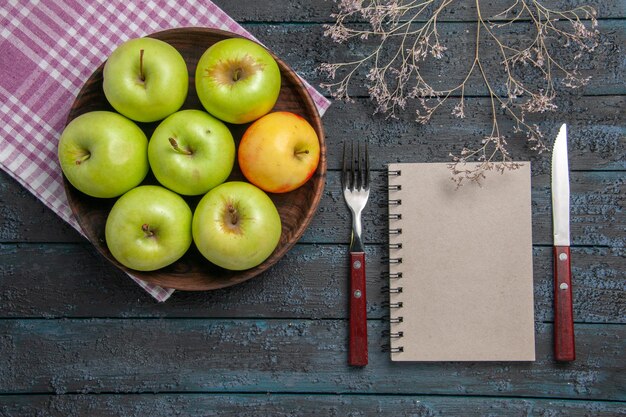 Vista superior del tazón de fuente de manzanas de siete manzanas de color amarillo verdoso sobre un mantel a cuadros junto a ramas, tenedor, cuchillo y cuaderno gris