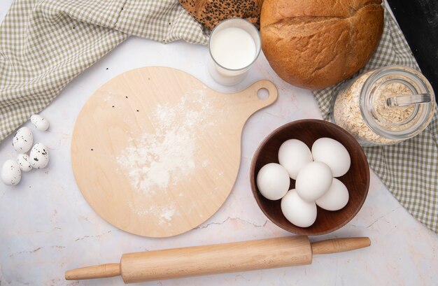 Vista superior del tazón de huevos y tabla de cortar con rodillo y leche copos de avena pan de mazorca sobre superficie blanca