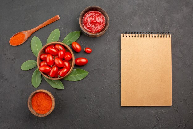 Vista superior de un tazón de laurel de tomates cherry deja una cuchara de madera y tazones de salsa de tomate y pimiento picante en polvo y un cuaderno sobre la mesa negra