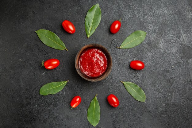 Vista superior de un tazón de salsa de tomate alrededor de tomates cherry y hojas de laurel sobre suelo oscuro