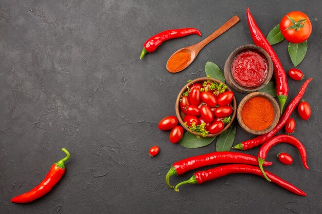 Vista superior de un tazón de tomates cherry pimientos rojos picantes una cuchara de madera hojas de laurel tazones de ketchup y pimiento picante en polvo y tomate sobre fondo negro