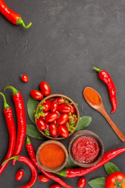 Vista superior de un tazón de tomates cherry, pimientos rojos picantes, una cuchara de madera, hojas de laurel, tazones de salsa de tomate y pimiento picante en polvo y tomate en el fondo de la tierra negra