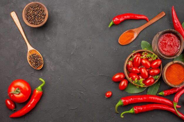 Vista superior de un tazón de tomates cherry pimientos rojos picantes pimienta negra en una cuchara de madera tazones de fuente de salsa de tomate pimienta negra y pimiento rojo en polvo sobre la mesa negra