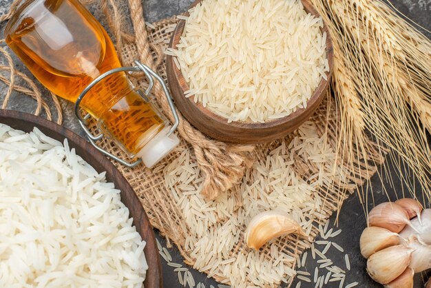 Vista superior de tazones largos perfectos de arroz marrón sobre una toalla de color desnudo, botellas de aceite caídas, picos de ajo sobre fondo oscuro