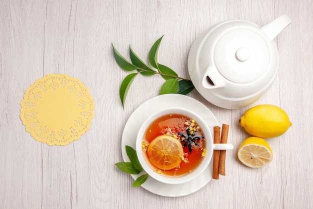 Vista superior de té y canela una taza de té de hierbas palitos de canela en el platillo junto a la tetera de limón tapete de encaje y hojas sobre la mesa blanca